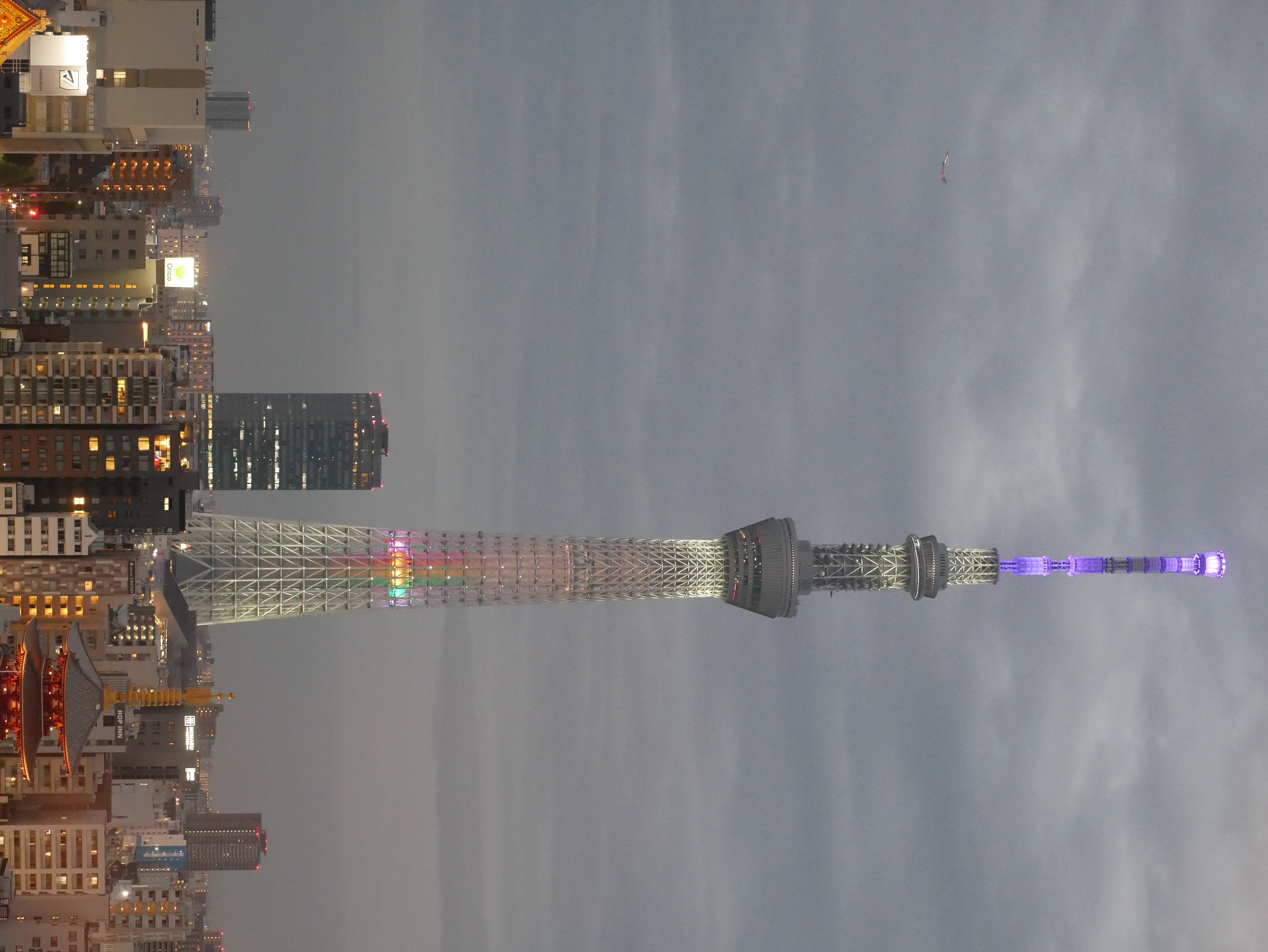 Tokyo Skytree, early evening. Dark skies, Sensō-ji temple lit, bright city lights and narrow streets. The Tokyo Skytree is a structure that starts with a base and thins out towards the top, has two observation decks two thirds of the way to the top and is lit in purple at the top and in rainbow LED colours across the tower.