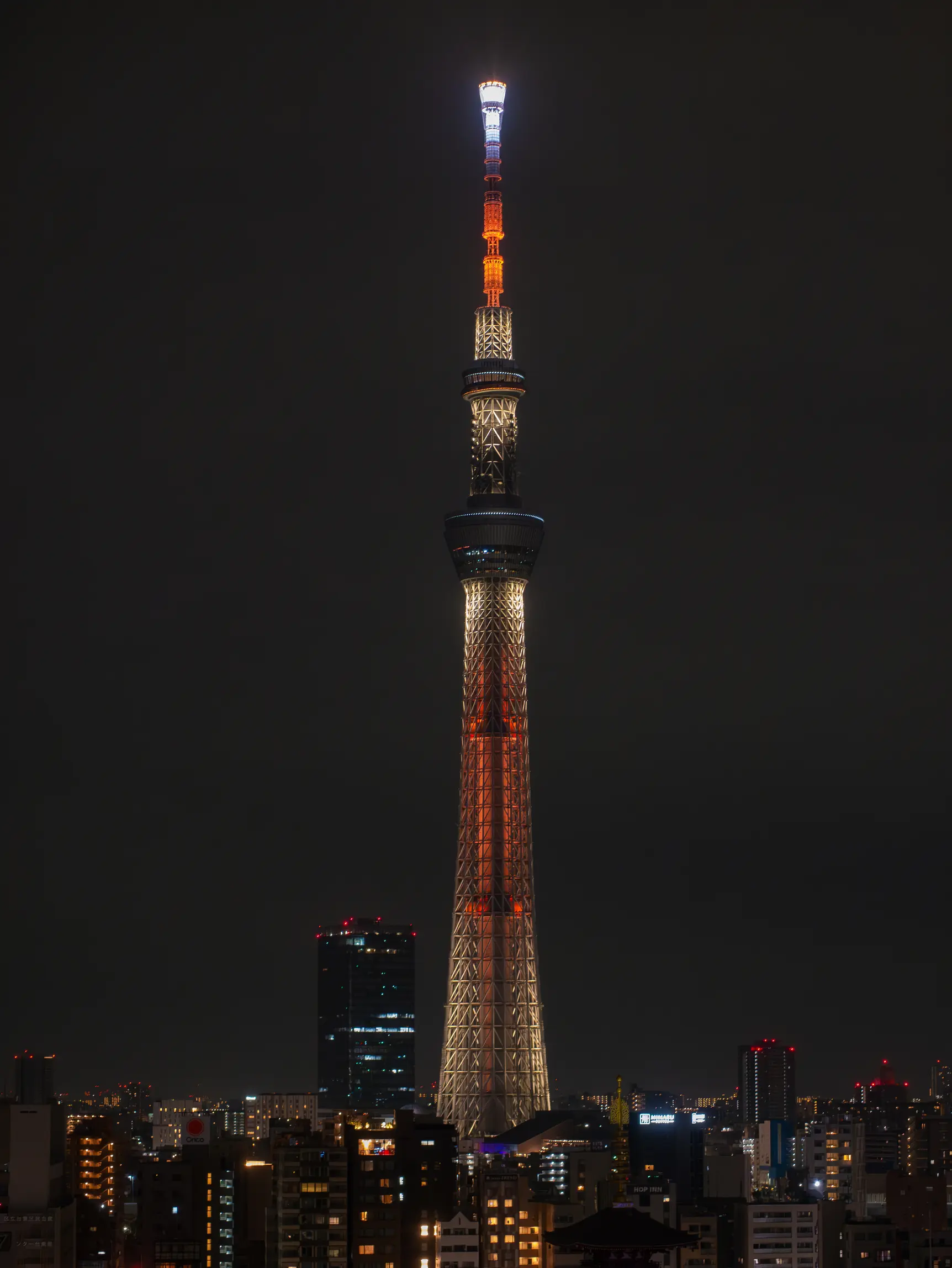 Tokyo Skytree, around 10:30 PM JST. Dark skies, warm city lights, narrow streets and Sensō-ji temple in view. The Tokyo Skytree is a structure that starts with a base and thins out towards the top, has two observation decks two thirds of the way to the top and is lit in red and white LED colours across the tower.