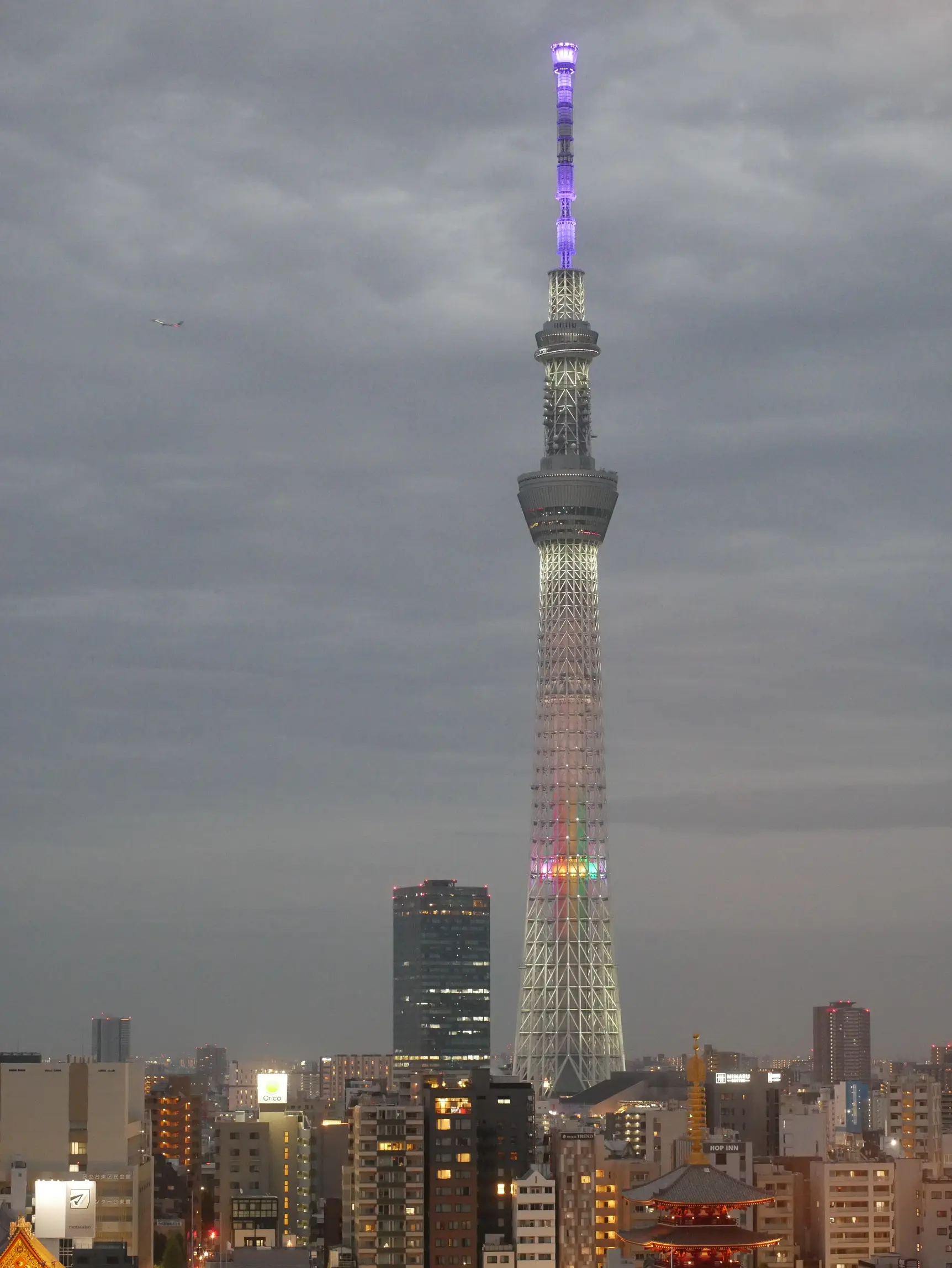 Tokyo Skytree, early evening. Dark skies, Sensō-ji temple lit, bright city lights and narrow streets. The Tokyo Skytree is a structure that starts with a base and thins out towards the top, has two observation decks two thirds of the way to the top and is lit in purple at the top and in rainbow LED colours across the tower.